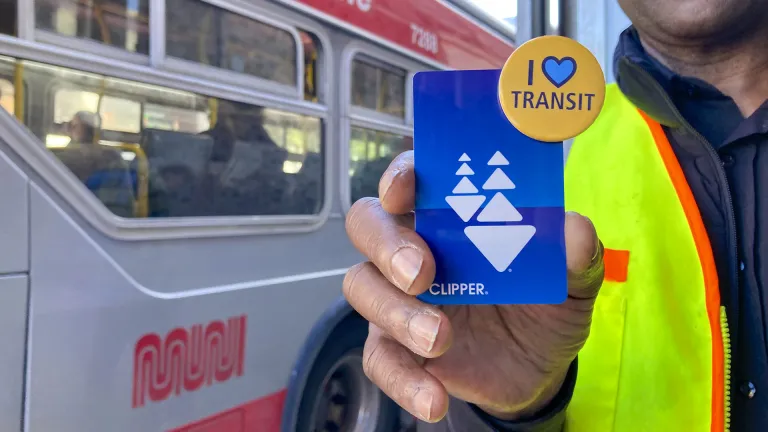 Closeup of a man's hand holding a Clipper card and an "I Heart Transit" pin, as a Muni bus passes by.