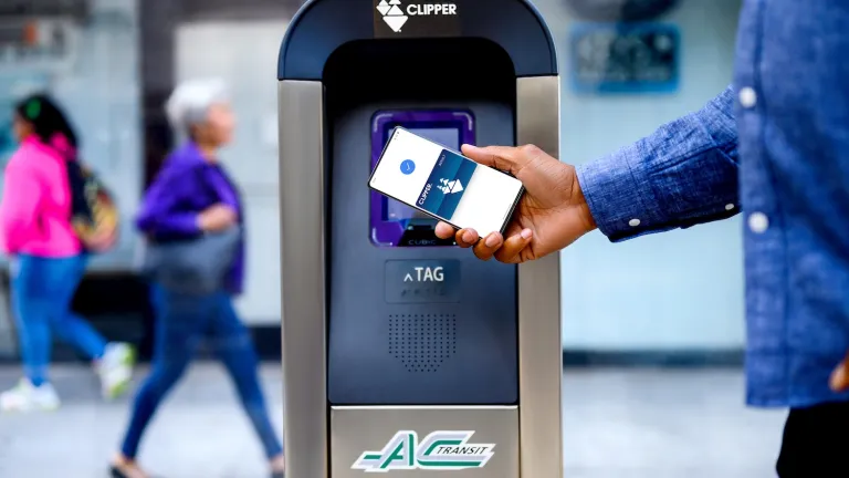 A hand holds a phone next to an AC Transit Clipper card reader with a mobile Clipper card displayed on the phone.