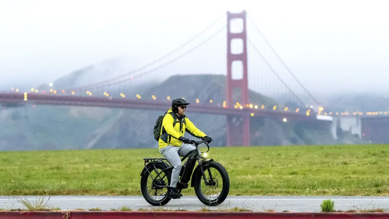 A cyclist on an e-bike rides through Crissy Field in San Francisco, with the fog-covered Golden Gate Bridge in the background.