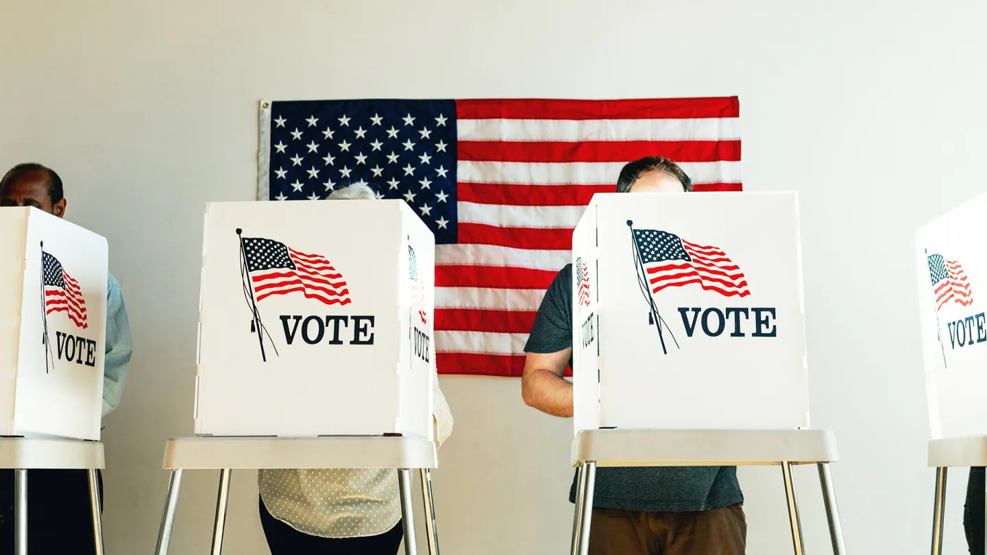 People at voting booths, with an American flag behind them.