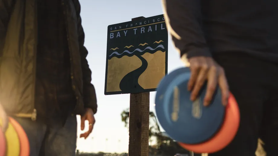 People holding Frisbee flying discs in front of a Bay Trail sign.