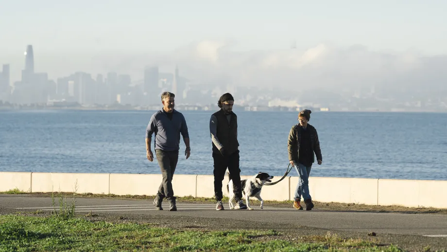 People walking a dog on the Bay Trail, with the foggy San Francisco skyline behind.