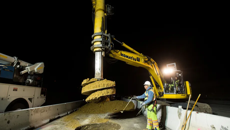 An excavator with a drill attachment pre-drills a shaft where metal rebar cages and concrete will be placed.