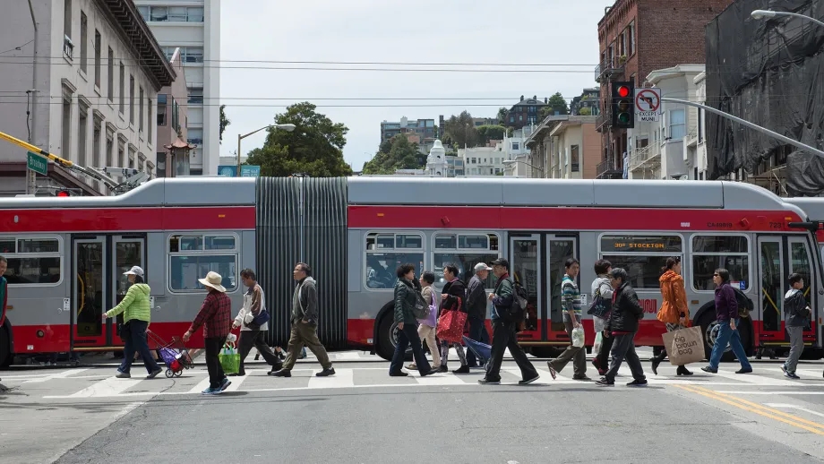 A crowd of pedestrians in a crosswalk as an articulated Muni bus passes by.