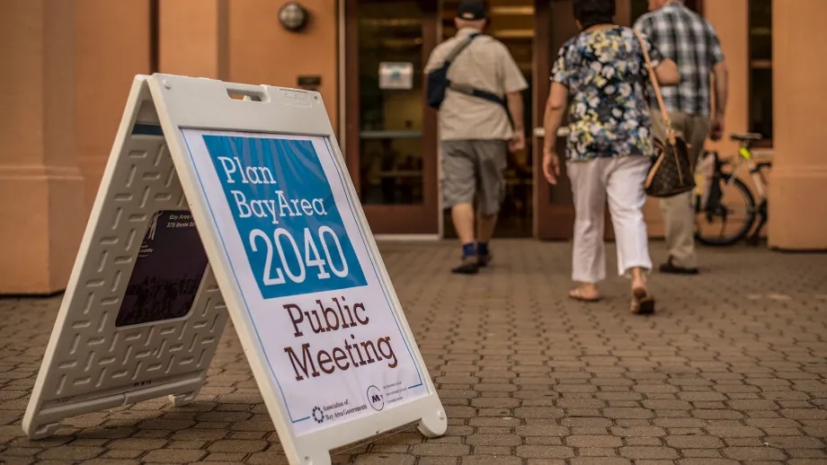 In the foreground a sandwich board advertising the Plan Bay Area 2040 public meeting is displayed, with three adults walking into the building in the background.