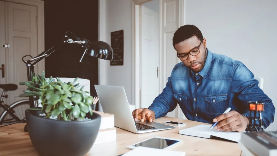 A man works at a laptop while writing in a notebook, with a bicycle in the background.