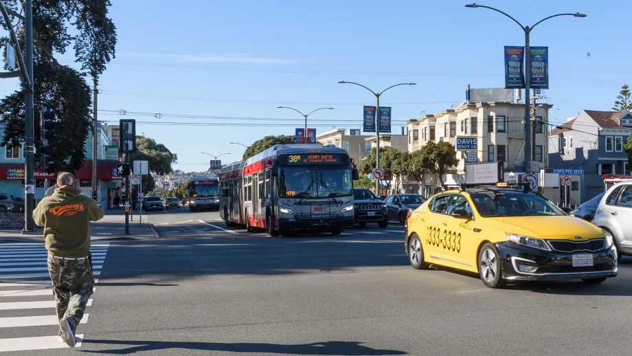 A pedestrian in a crosswalk, a taxi, and a Muni bus at an intersection in San Francisco.